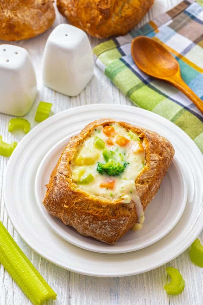 A bread bowl filled with creamy vegetable chowder, topped with broccoli and carrots, perfect for serving a cozy meal.