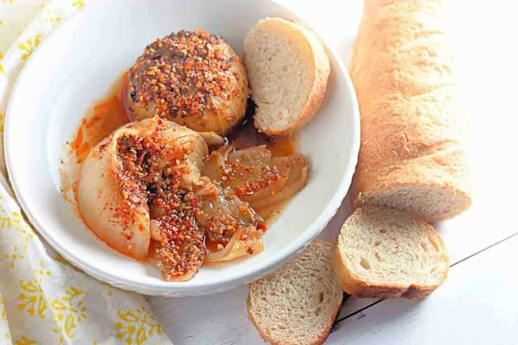 Baked stuffed onions in a reddish sauce, seasoned with spices and served in a white bowl, surrounded by crusty bread and a baguette.
