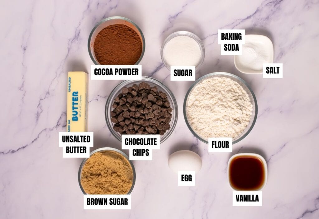 Baking ingredients laid out on a marble surface, labeled bowls of cocoa powder, sugar, flour, baking soda, salt, chocolate chips, butter, egg, brown sugar, and vanilla.