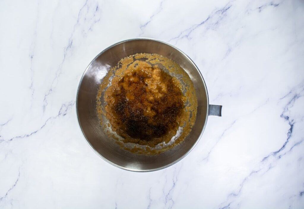 A close-up of a mixing bowl containing a mixture of browned butter and sugars, with a slight bubbly texture.