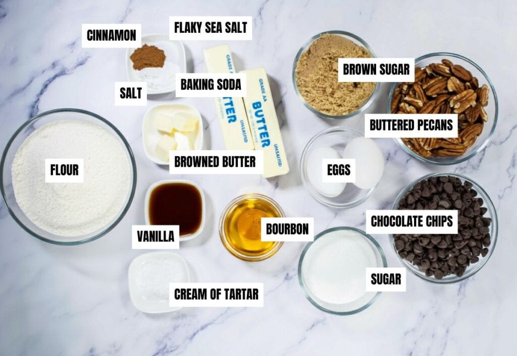 A neatly arranged collection of ingredients for making browned butter bourbon pecan chocolate chip cookies, including butter, flour, chocolate chips, pecans, and spices.