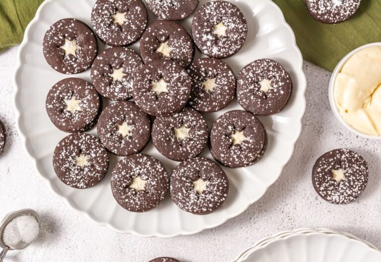 A plate of chocolate Linzer cookies with cream cheese filling and a dusting of powdered sugar, arranged neatly in a circular pattern.