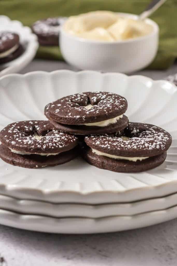 A plate of chocolate Linzer cookies with cream cheese filling and powdered sugar dusting, arranged in a neat stack with a bowl of frosting in the background.