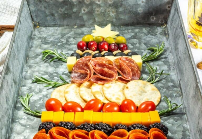 A festive charcuterie board arranged in the shape of a Christmas tree, featuring slices of salami, crackers, cheese, olives, rosemary, cherry tomatoes, and dried fruits like cranberries and blackberries, topped with a cheese star.