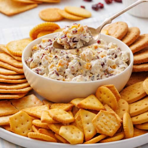 A close-up shot of a creamy cranberry dip surrounded by a variety of crackers on a white plate with a spoonful of dip being lifted.