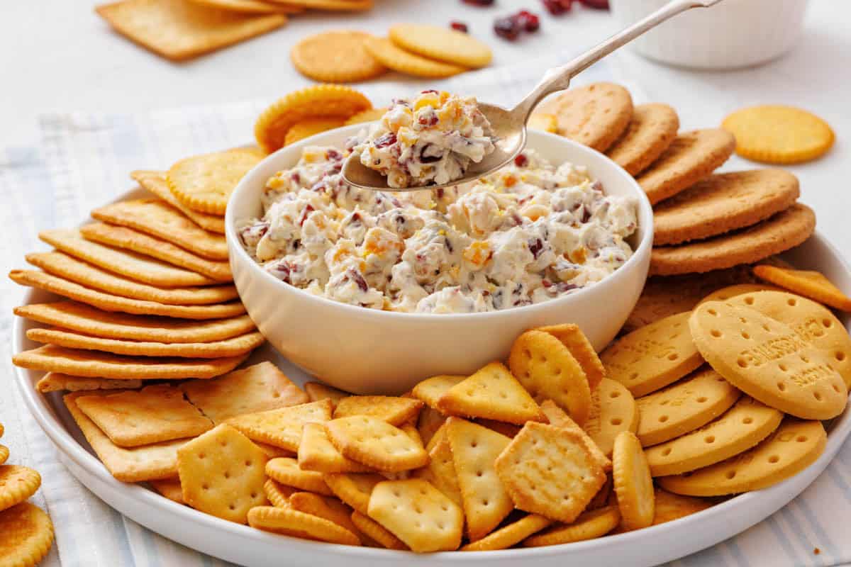A close-up shot of a creamy cranberry dip surrounded by a variety of crackers on a white plate with a spoonful of dip being lifted.