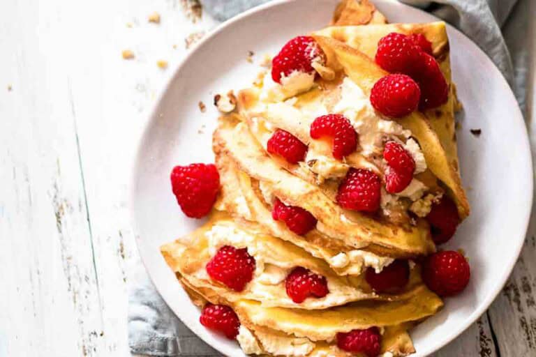 A plate of crepes filled with cream cheese and topped with fresh raspberries, nuts, and a light dusting of powdered sugar.