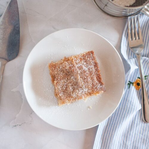 A slice of gooey butter cake dusted with powdered sugar, served on a white plate with a fork beside it.
