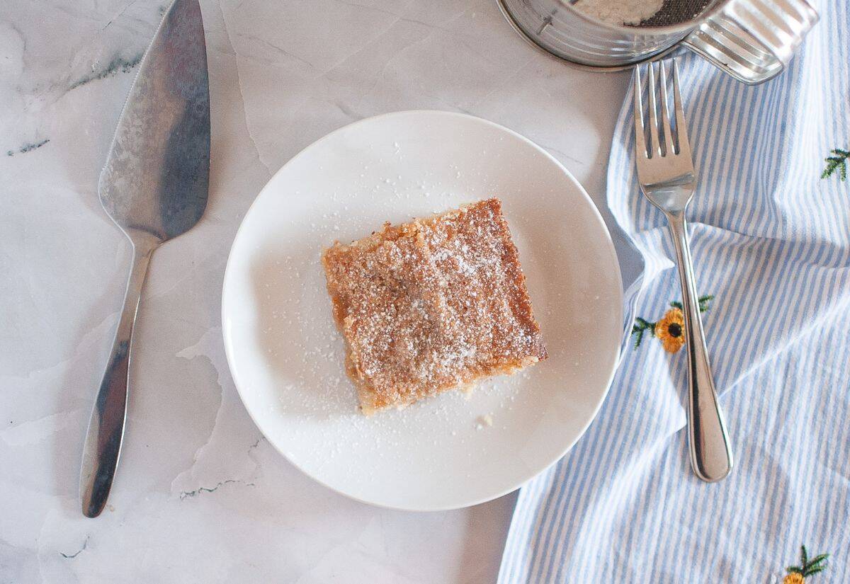 A slice of gooey butter cake dusted with powdered sugar, served on a white plate with a fork beside it.