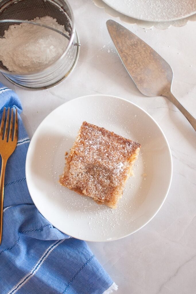 A slice of gooey butter cake on a plate with powdered sugar sprinkled on top, with a cake server and fork nearby.