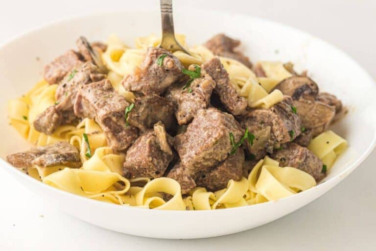 A white bowl filled with cooked egg noodles and chunks of beef, garnished with herbs, with a fork standing upright in the center.