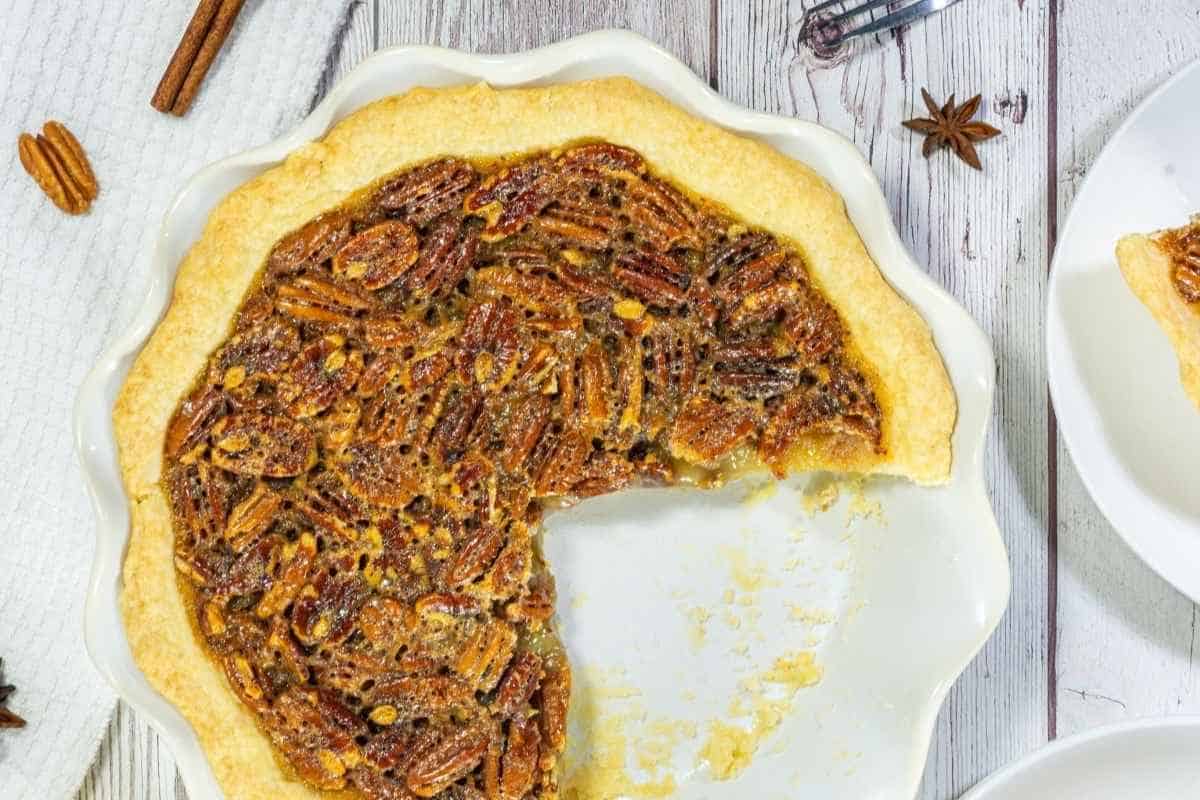 A pecan pie in a white dish with a large slice missing, shown on a light wooden surface.