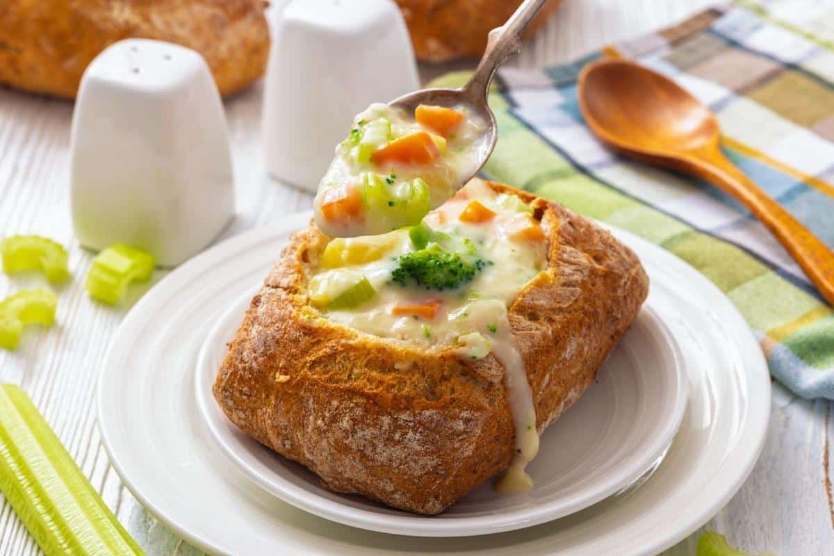 A bread bowl filled with creamy vegetable chowder, with a spoon scooping soup out; salt and pepper shakers, celery, and a wooden spoon are nearby.