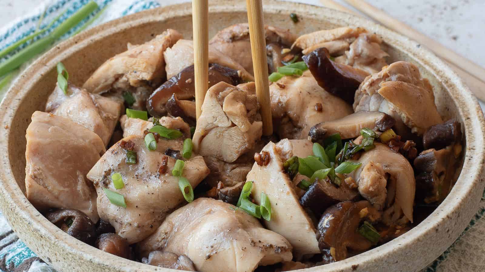 A bowl of steamed chicken pieces with mushrooms, garnished with chopped green onions, with chopsticks picking up a piece.