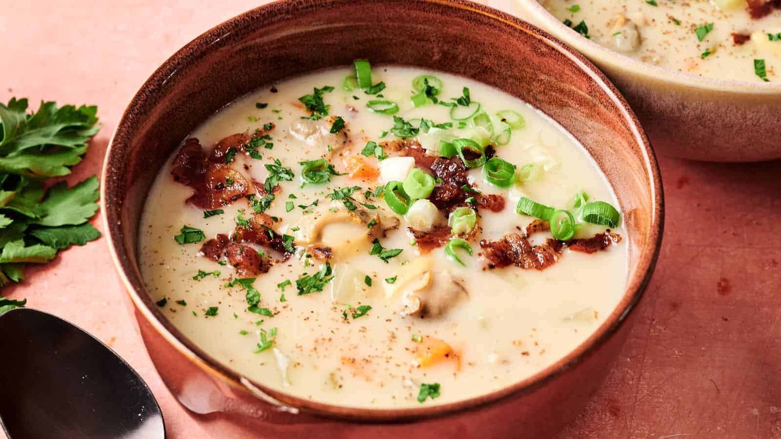A bowl of creamy soup with clams, bacon, chopped green onions, and parsley, placed on a pink surface next to a spoon and some fresh herbs.