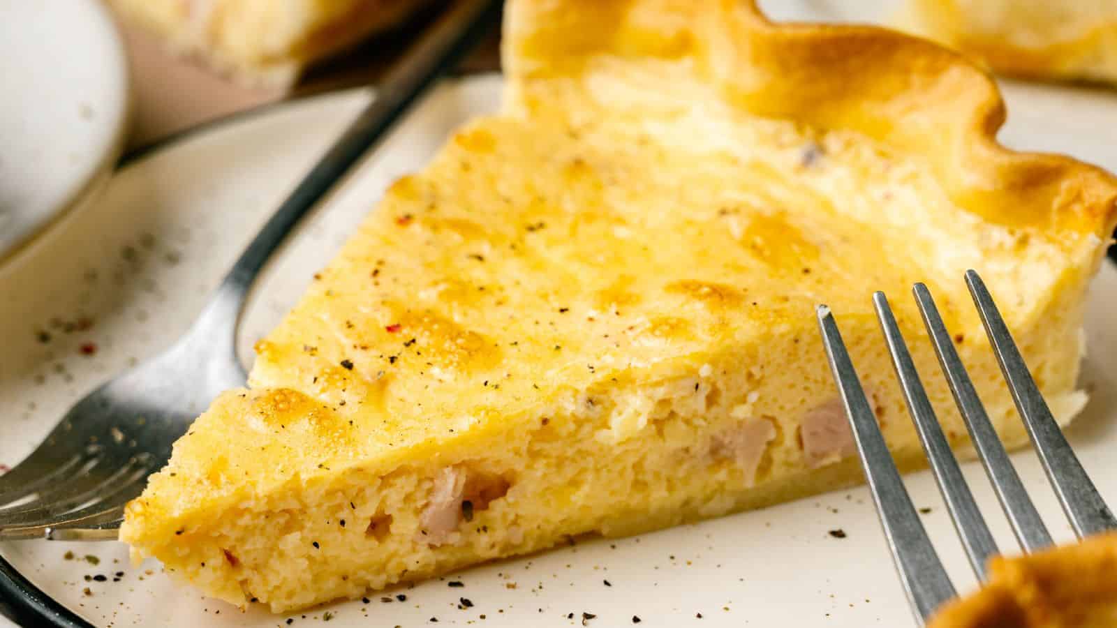 A close-up of a slice of quiche on a plate, with a fork beside it. The quiche has a golden crust and a creamy, egg-filled interior.