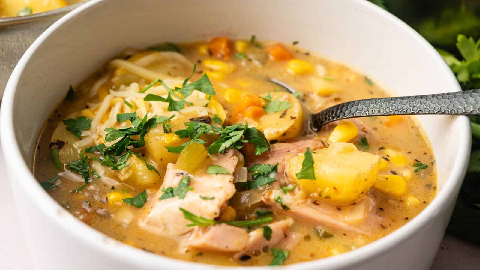 A bowl of creamy chicken and vegetable soup with potatoes, corn, carrots, and fresh parsley, served with a metal spoon.