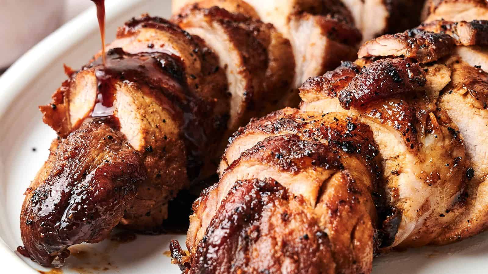 Close-up of roasted pork tenderloin on a white plate, topped with a drizzle of dark sauce. The meat is seasoned with visible spices and has a textured, caramelized surface.
