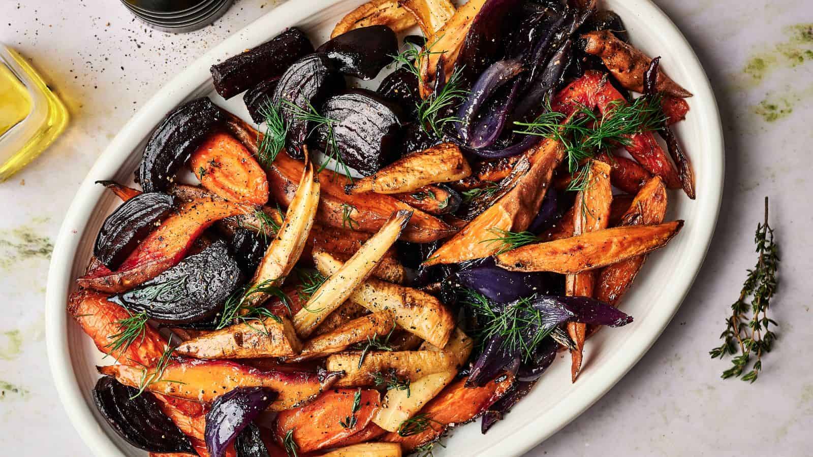 A white dish with roasted root vegetables, including carrots, parsnips, red onions, and sweet potatoes, garnished with fresh dill and thyme.