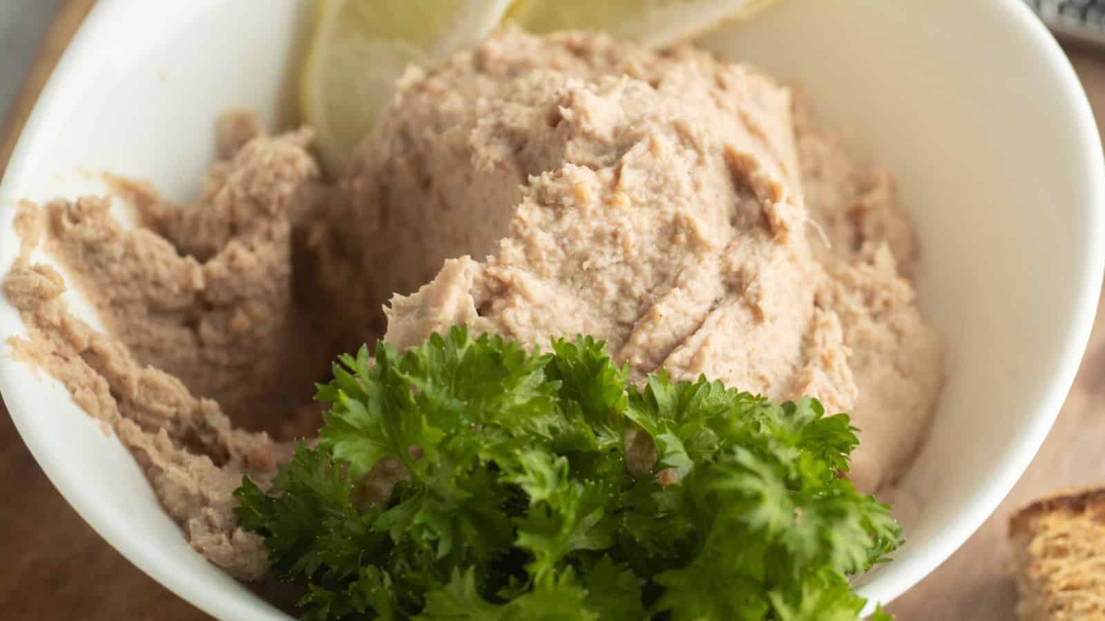 A white bowl filled with a creamy, light brown spread, garnished with fresh parsley and lemon slices.