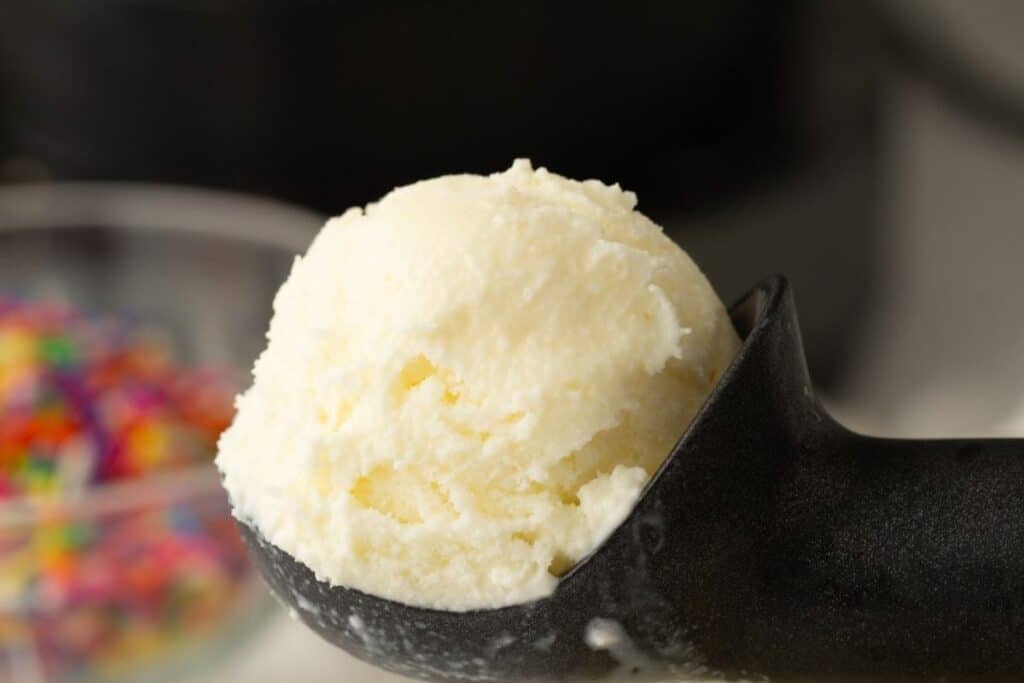 A close-up of a scoop of vanilla ice cream in a black ice cream scooper, with colorful sprinkles blurred in the background.