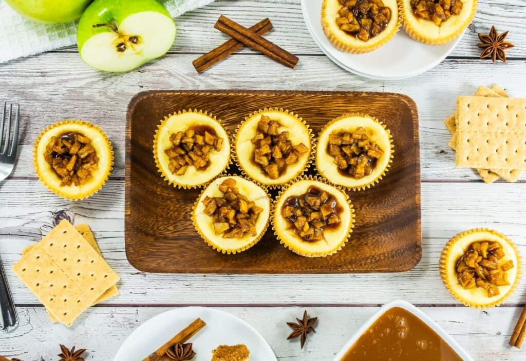 A wooden platter with mini caramel apple cheesecakes topped with spiced caramel apples, surrounded by graham crackers, cinnamon sticks, and fresh Granny Smith apples.