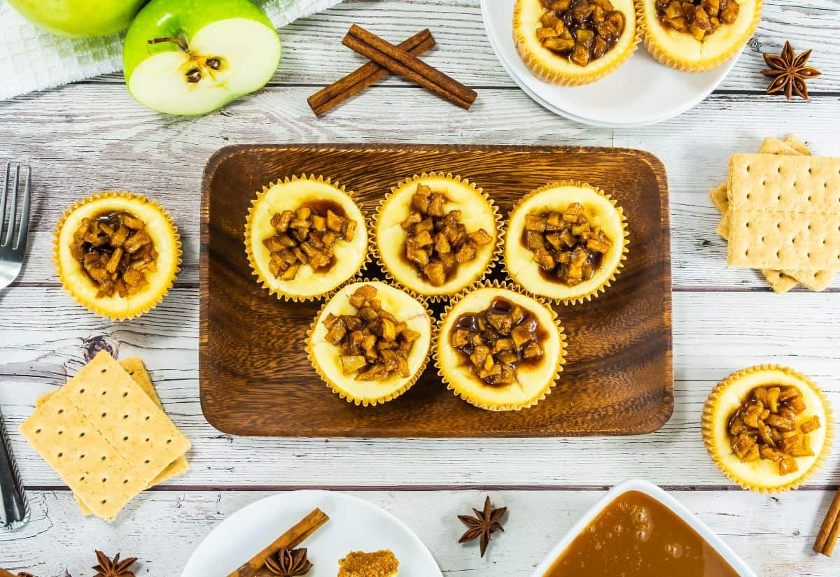 A wooden platter with mini caramel apple cheesecakes topped with spiced caramel apples, surrounded by graham crackers, cinnamon sticks, and fresh Granny Smith apples.