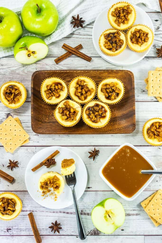 A close-up of a mini caramel apple cheesecake with a fork taking a bite, revealing the creamy filling and golden graham cracker crust, topped with caramel apple topping.