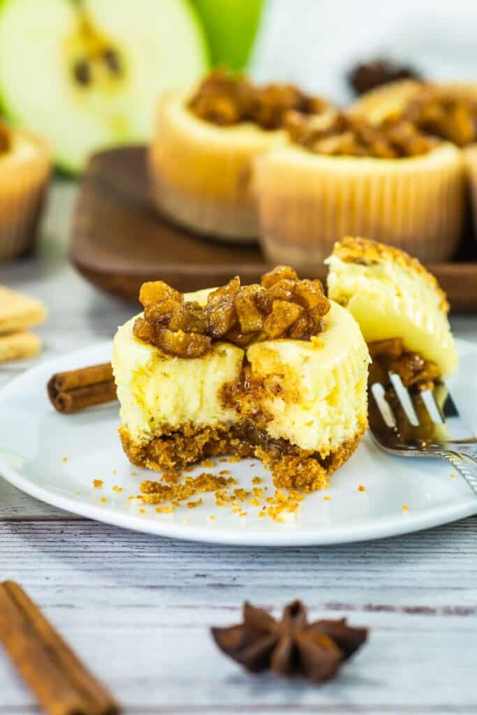A close-up of a platter of mini caramel apple cheesecakes with a spoonful of caramel apple topping, accompanied by cinnamon sticks, star anise, and fresh apple slices.