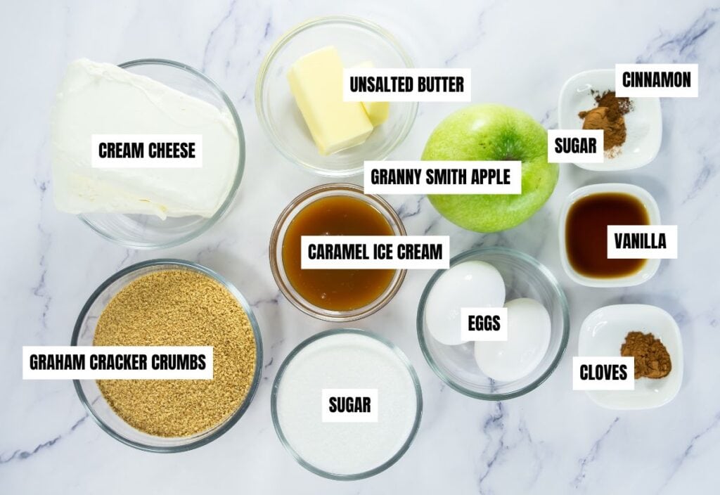 A set of ingredients for mini caramel apple cheesecakes, including cream cheese, unsalted butter, a Granny Smith apple, caramel ice cream topping, cinnamon, sugar, eggs, graham cracker crumbs, and cloves.
