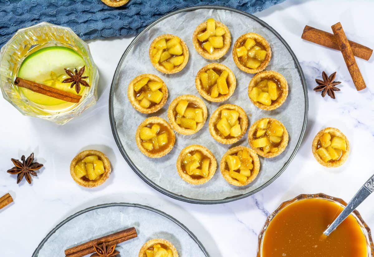 A close-up shot of mini caramel apple tartlets on a plate, garnished with cinnamon sticks and star anise, creating a cozy, autumnal vibe.