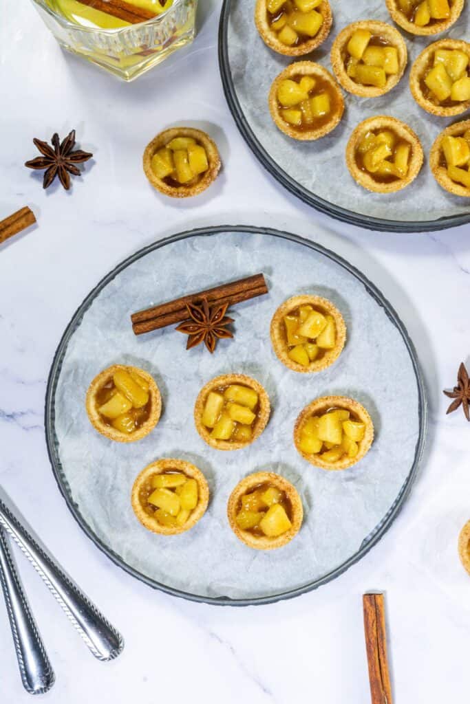 A close-up of a few mini caramel apple tartlets placed on parchment paper, with cinnamon and star anise accents in the background.