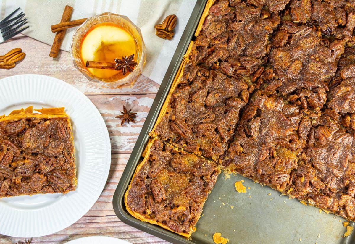 A close-up of a pecan slab pie served with a slice on a plate, accompanied by a cinnamon stick drink and scattered pecans and spices.