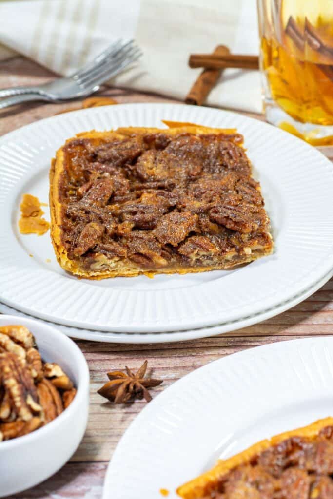 A close-up of a slice of pecan slab pie on a plate, with scattered pecans and a cinnamon stick drink in the background.