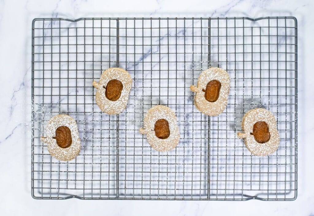 Assembled Pumpkin Linzer Cookies, with powdered sugar dusted on top and pumpkin butter in the center, displayed on a wire rack.