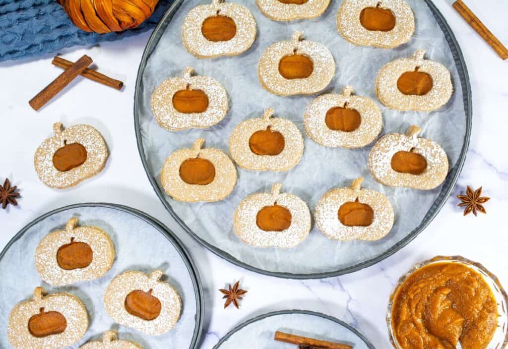 A plate of Pumpkin Linzer Cookies dusted with powdered sugar, featuring a pumpkin-shaped design with a pumpkin butter center and surrounded by cinnamon sticks and star anise.