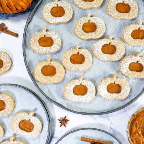 A plate of Pumpkin Linzer Cookies dusted with powdered sugar, featuring a pumpkin-shaped design with a pumpkin butter center and surrounded by cinnamon sticks and star anise.