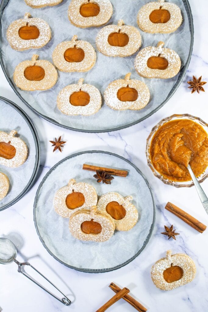 A close-up of Pumpkin Linzer Cookies with pumpkin-shaped cut-outs, dusted with powdered sugar and arranged on a tray with cinnamon sticks and star anise nearby.