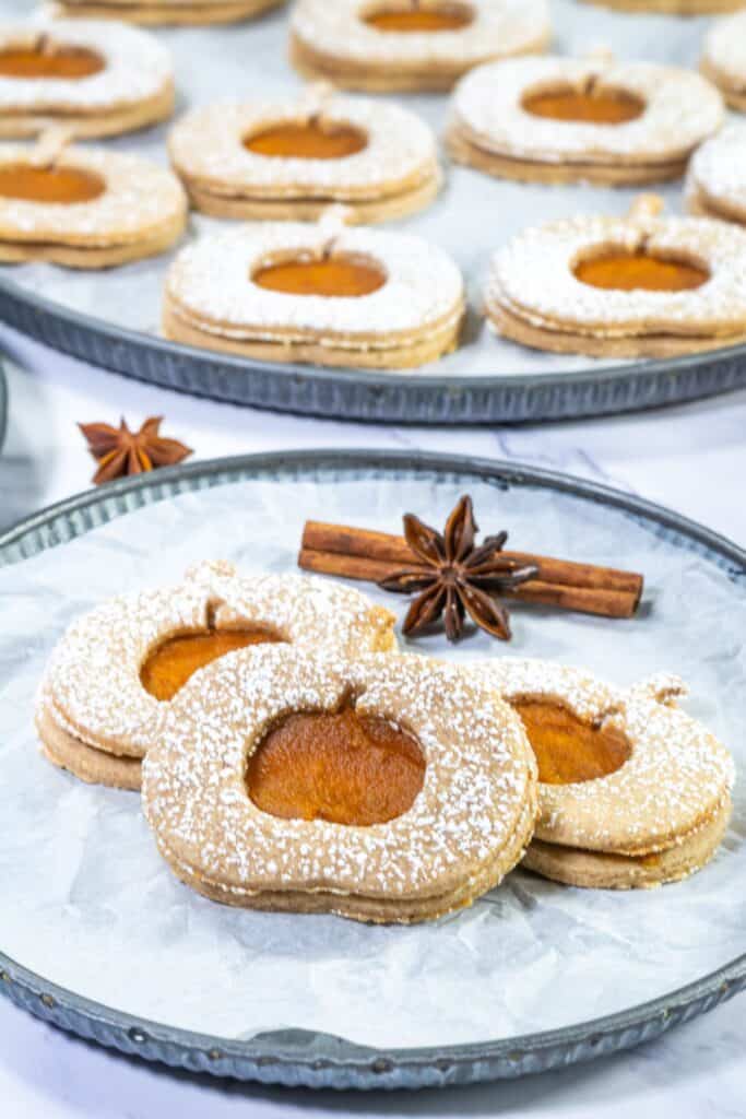 A tray of Pumpkin Linzer Cookies with pumpkin-shaped centers, dusted with powdered sugar, placed on parchment paper, and surrounded by cinnamon sticks and star anise.