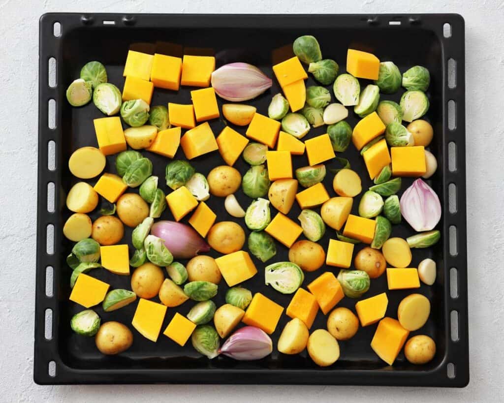 Overhead shot of a black baking sheet filled with raw, chopped butternut squash, Brussels sprouts, baby potatoes, and shallots, ready for roasting.