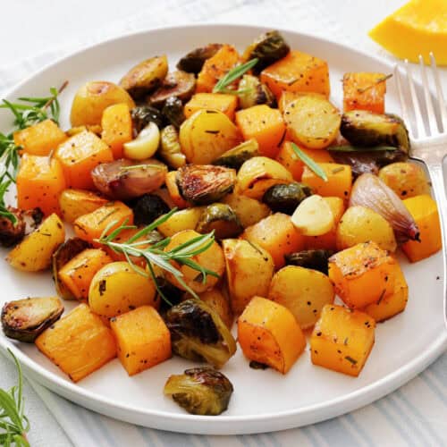 Plate of roasted butternut squash, Brussels sprouts, baby potatoes, garlic, and shallots garnished with fresh rosemary.