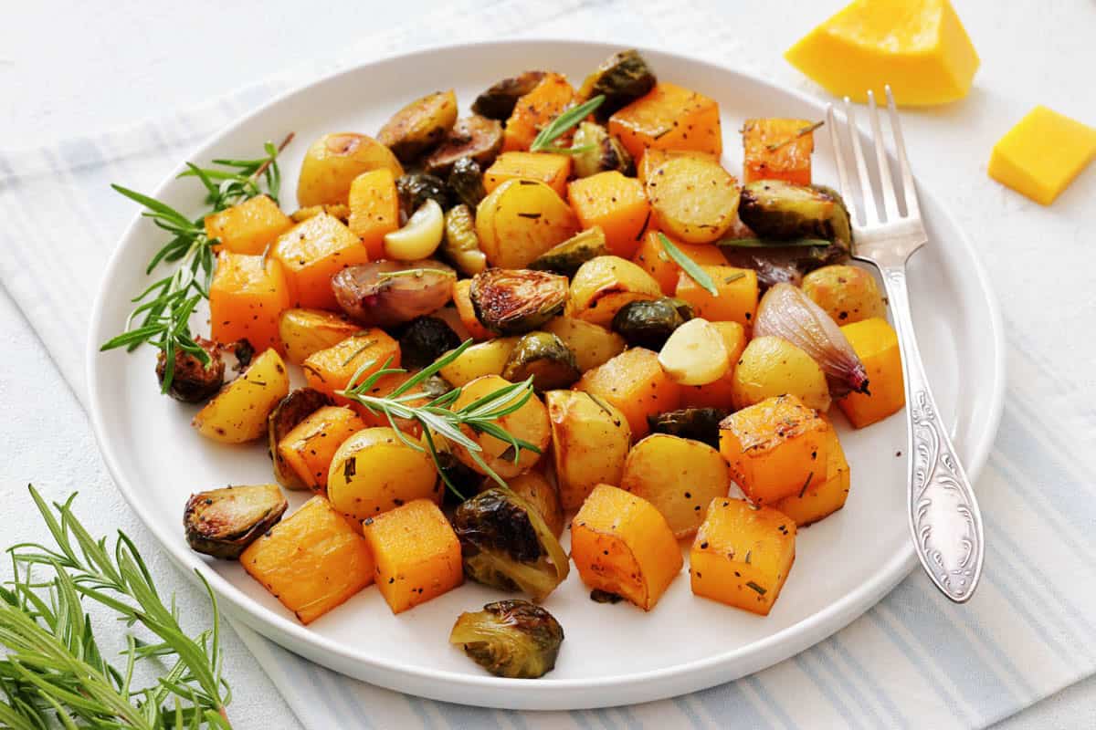 Plate of roasted butternut squash, Brussels sprouts, baby potatoes, garlic, and shallots garnished with fresh rosemary.