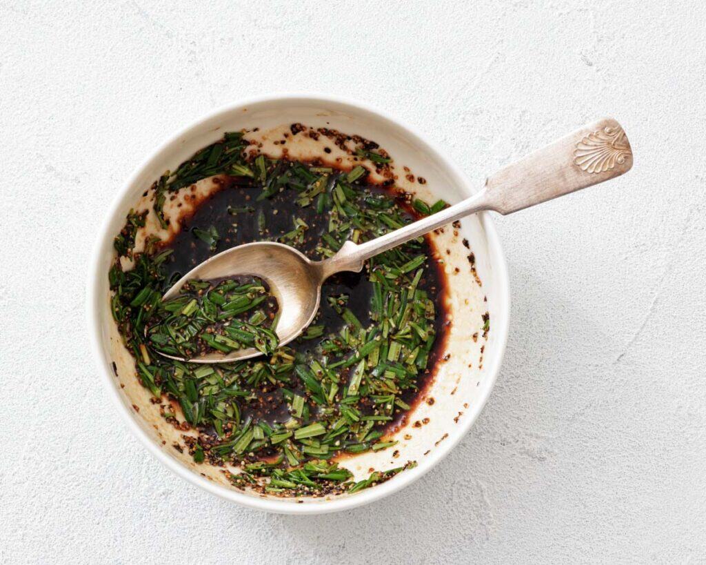 Overhead view of a small white bowl filled with dark brown vinaigrette, finely chopped green chives, and mustard seeds, a silver spoon rests within the bowl.