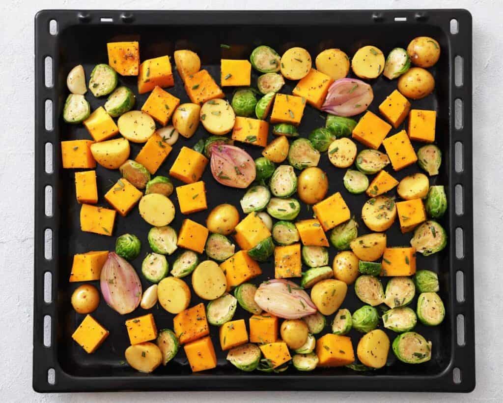 Roasted vegetables on a black baking sheet, including cubed butternut squash, halved baby potatoes, Brussels sprouts, halved shallots, and whole garlic cloves, all seasoned with herbs and spices.