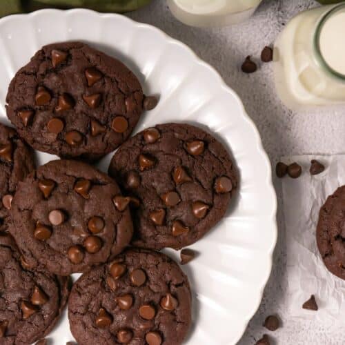 Top-down view of chocolate cookies on a white plate with chocolate chips, surrounded by milk and scattered chips.