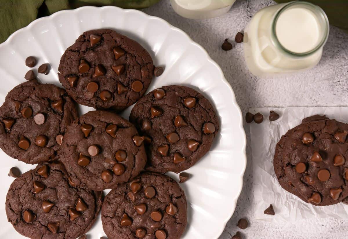 Top-down view of chocolate cookies on a white plate with chocolate chips, surrounded by milk and scattered chips.