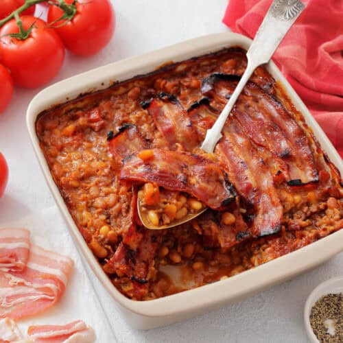 A casserole dish filled with Southern Ground Beef & Baked Bean Casserole topped with crispy bacon, placed next to fresh tomatoes and a red cloth.