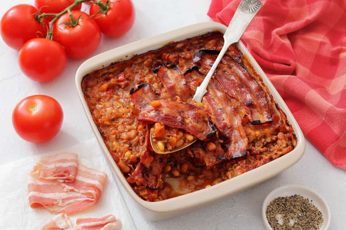 A casserole dish filled with Southern Ground Beef & Baked Bean Casserole topped with crispy bacon, placed next to fresh tomatoes and a red cloth.