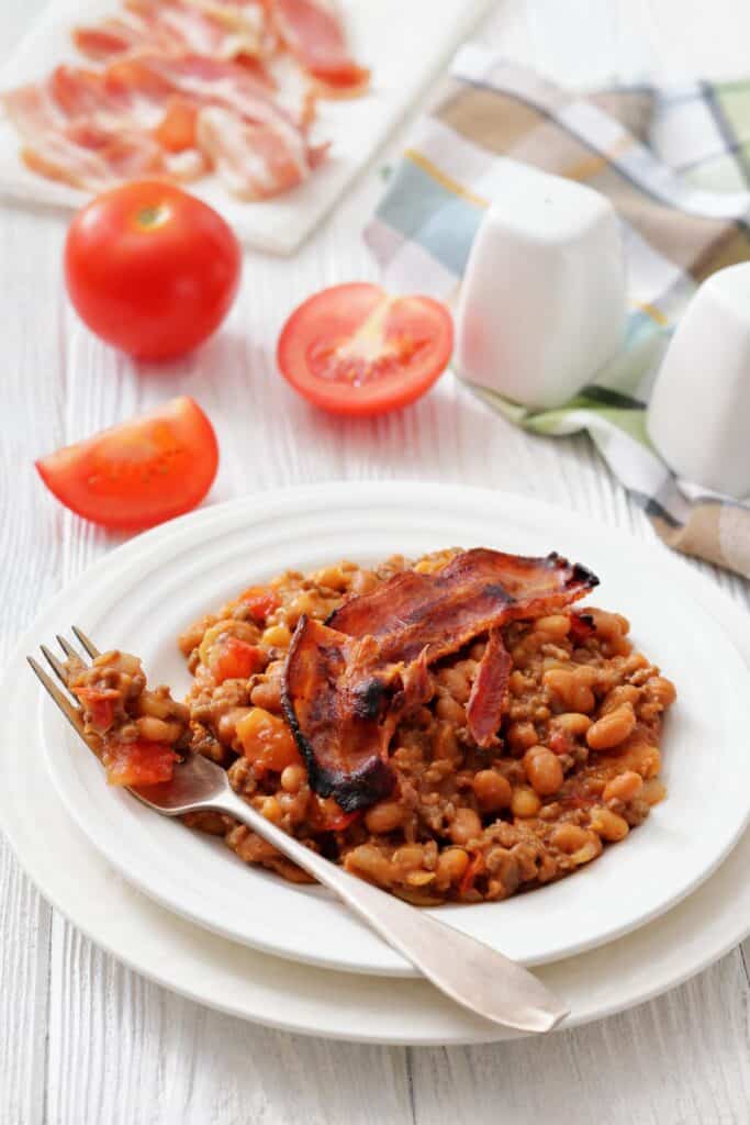 A close-up of Southern Ground Beef & Baked Bean Casserole with bacon, served on a plate next to fresh tomato slices.