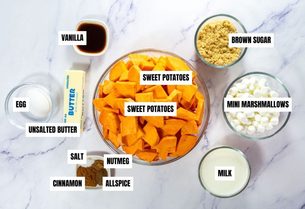 Overhead view of sweet potato casserole ingredients including chopped sweet potatoes, brown sugar, mini marshmallows, milk, butter, egg, vanilla, cinnamon, nutmeg, allspice, and salt on a marble surface.